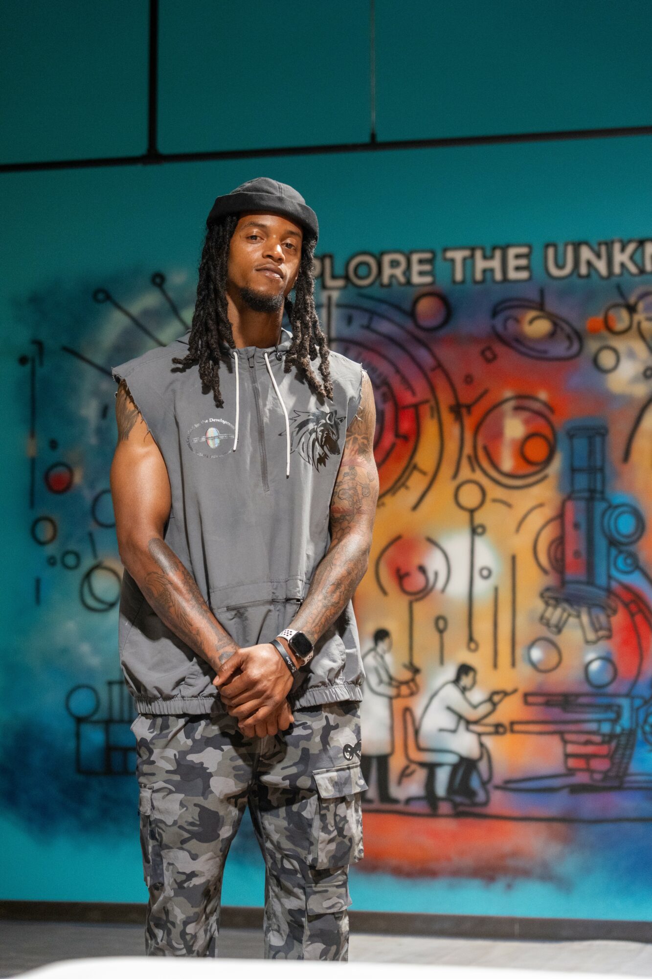 Young man with dreadlocks wearing a sleeveless hoodie and camouflage pants stands in front of a colorful mural.