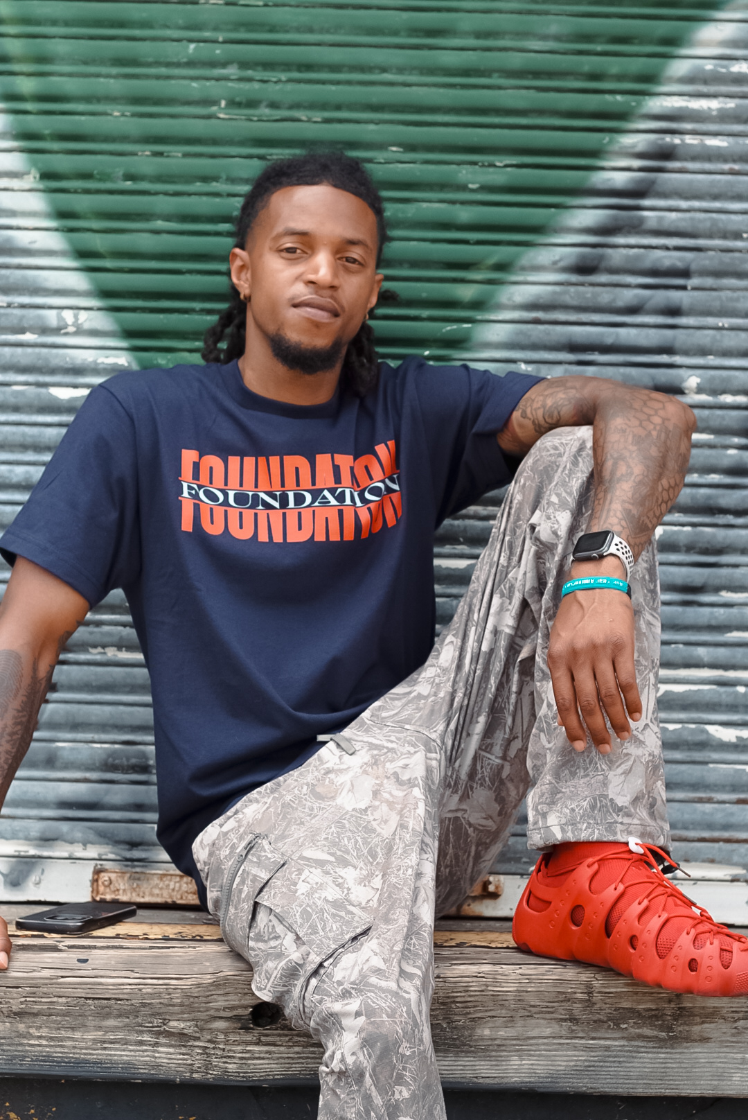 Man with dreadlocks sitting against a graffiti-covered wall, wearing a navy T-shirt, camouflage pants, and red Crocs, with a relaxed expression.