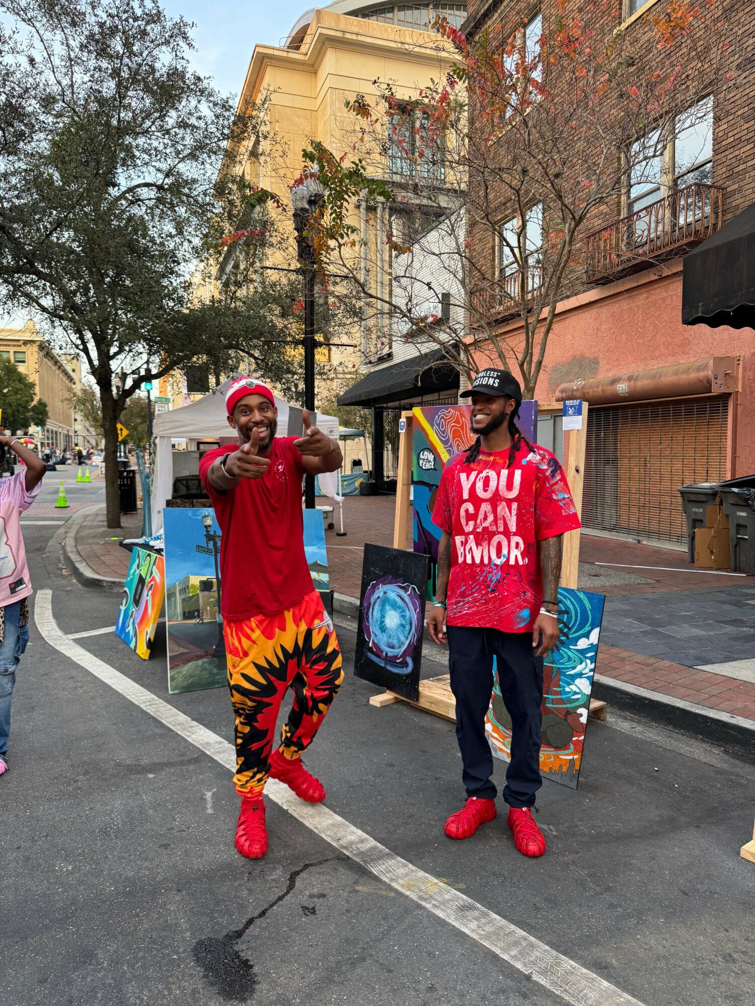 Two people in red costumes, one with a flame design, stand on a city street with buildings and trees in the background.