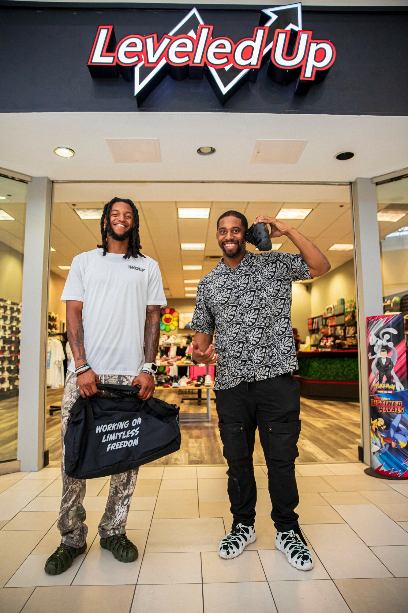 Two men standing in front of a store entrance with a sign reading 'Levelled Up.' One holds a bag, the other waves, both smiling.