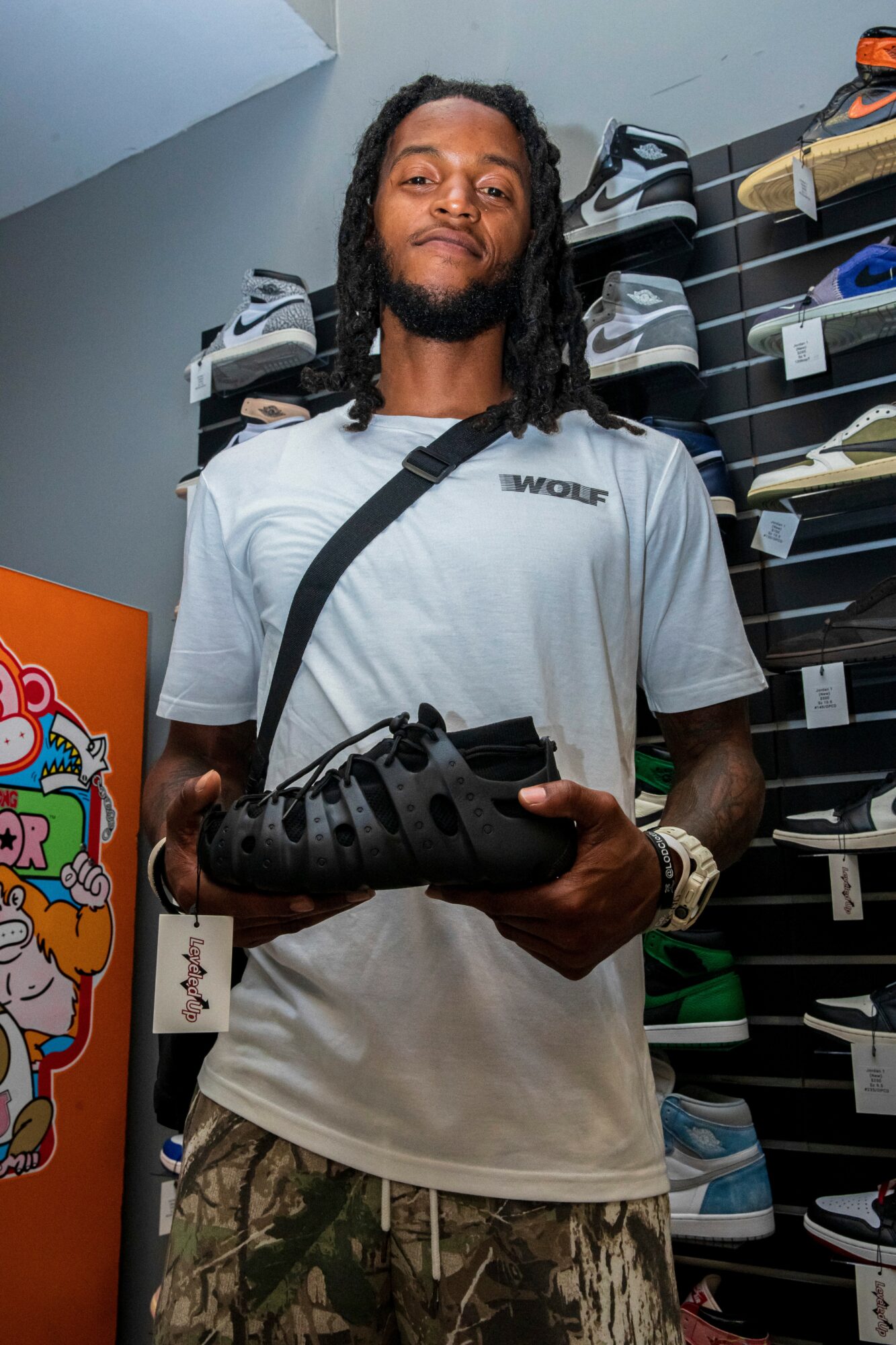 Young man with dreadlocks wearing a white t-shirt and camouflage shorts, holding a black sandal, standing in a store aisle.