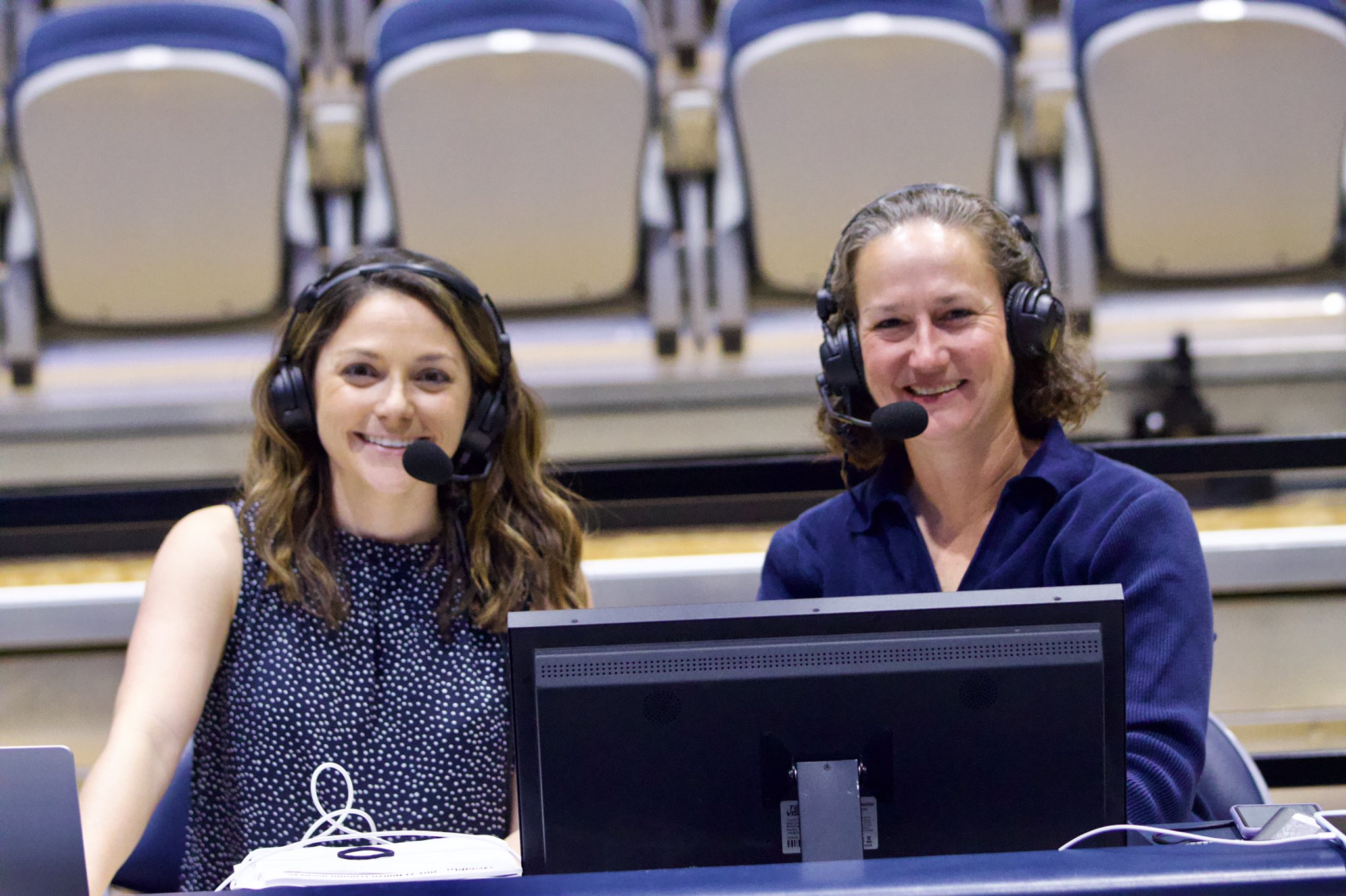 Two women with headsets sit behind a computer monitor, smiling, in a conference or broadcast setting.