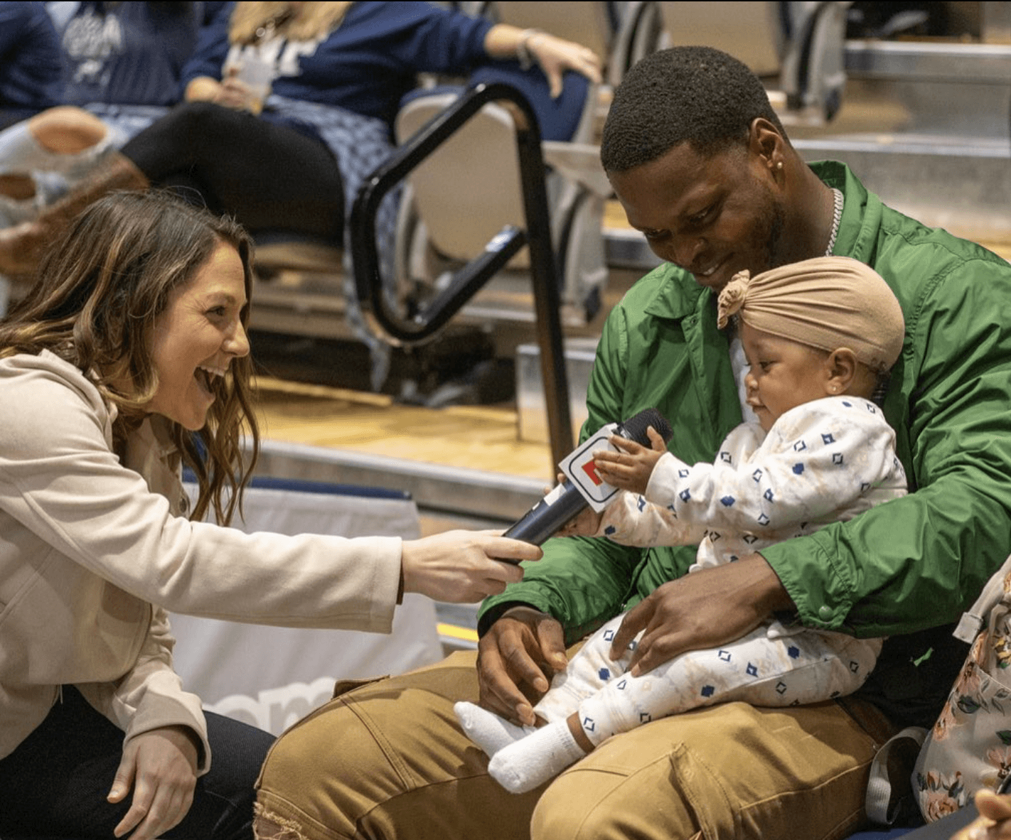 A woman interviews a man holding a baby in a public setting, with people and chairs in the background.