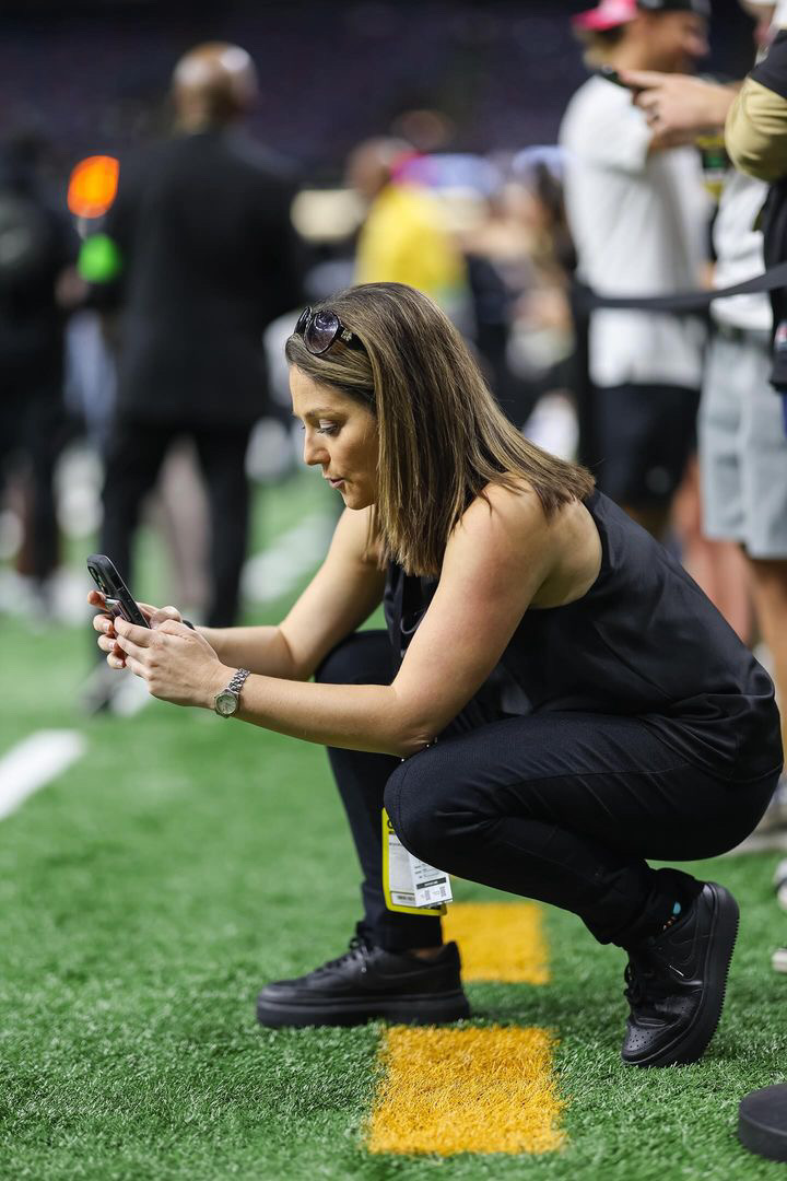 Woman crouching on grass, looking at her phone, with people standing in the background.