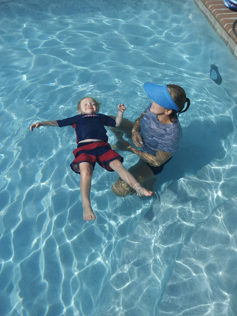 Child floating on back in swimming pool with adult holding and supporting them, sunlight reflecting on water surface.