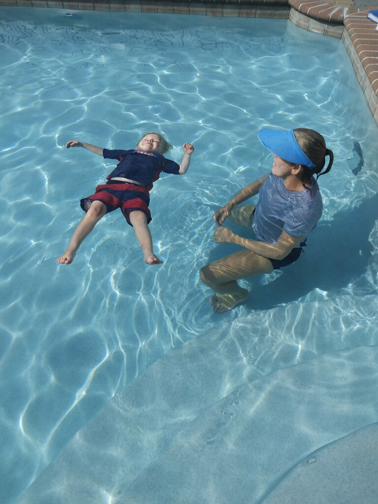 Child floating on back in swimming pool with adult sitting nearby, both in water, brick edge visible.