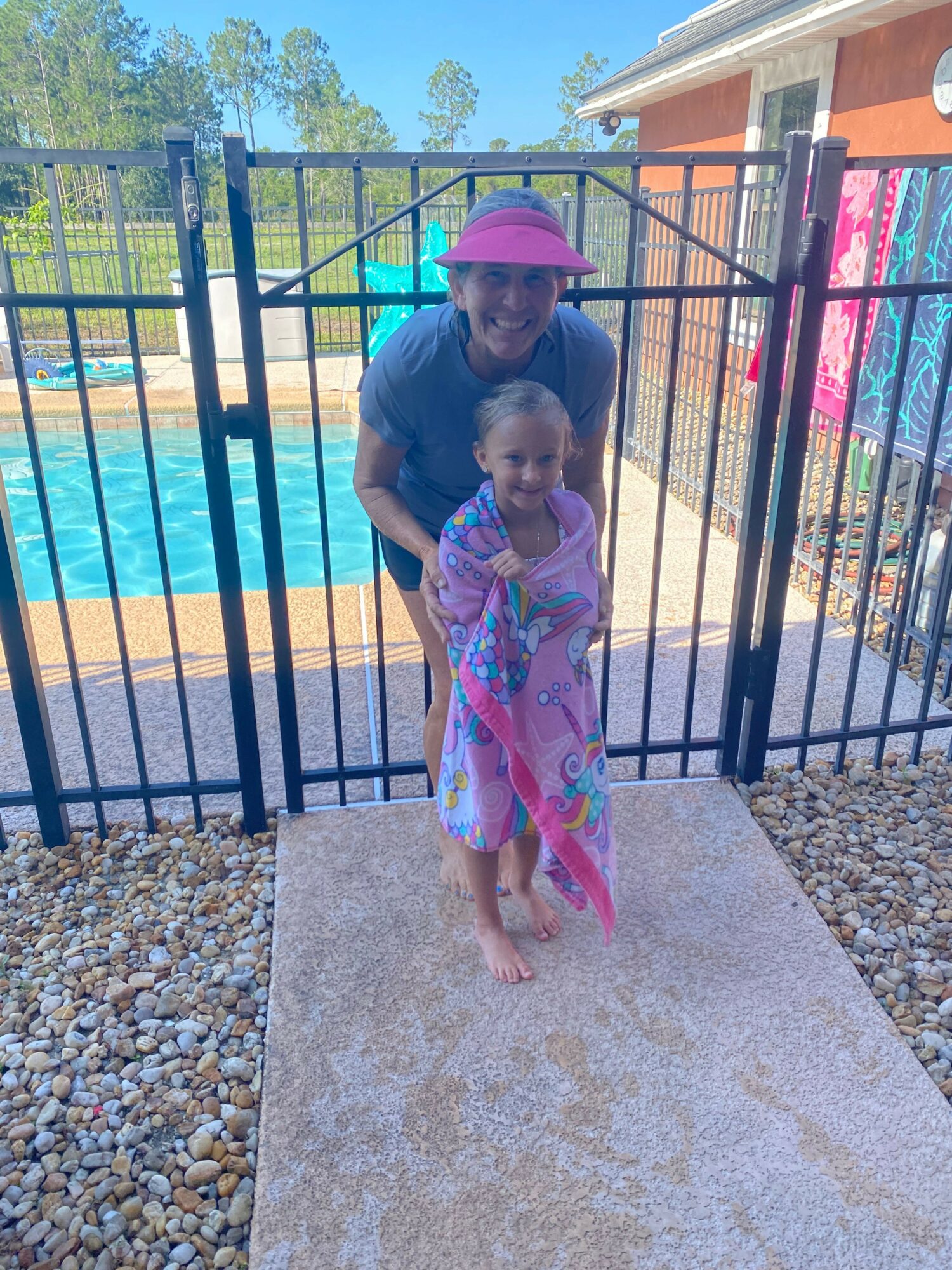 Child in colorful dress and hat standing on a concrete path near a pool, with a fence and trees in background.