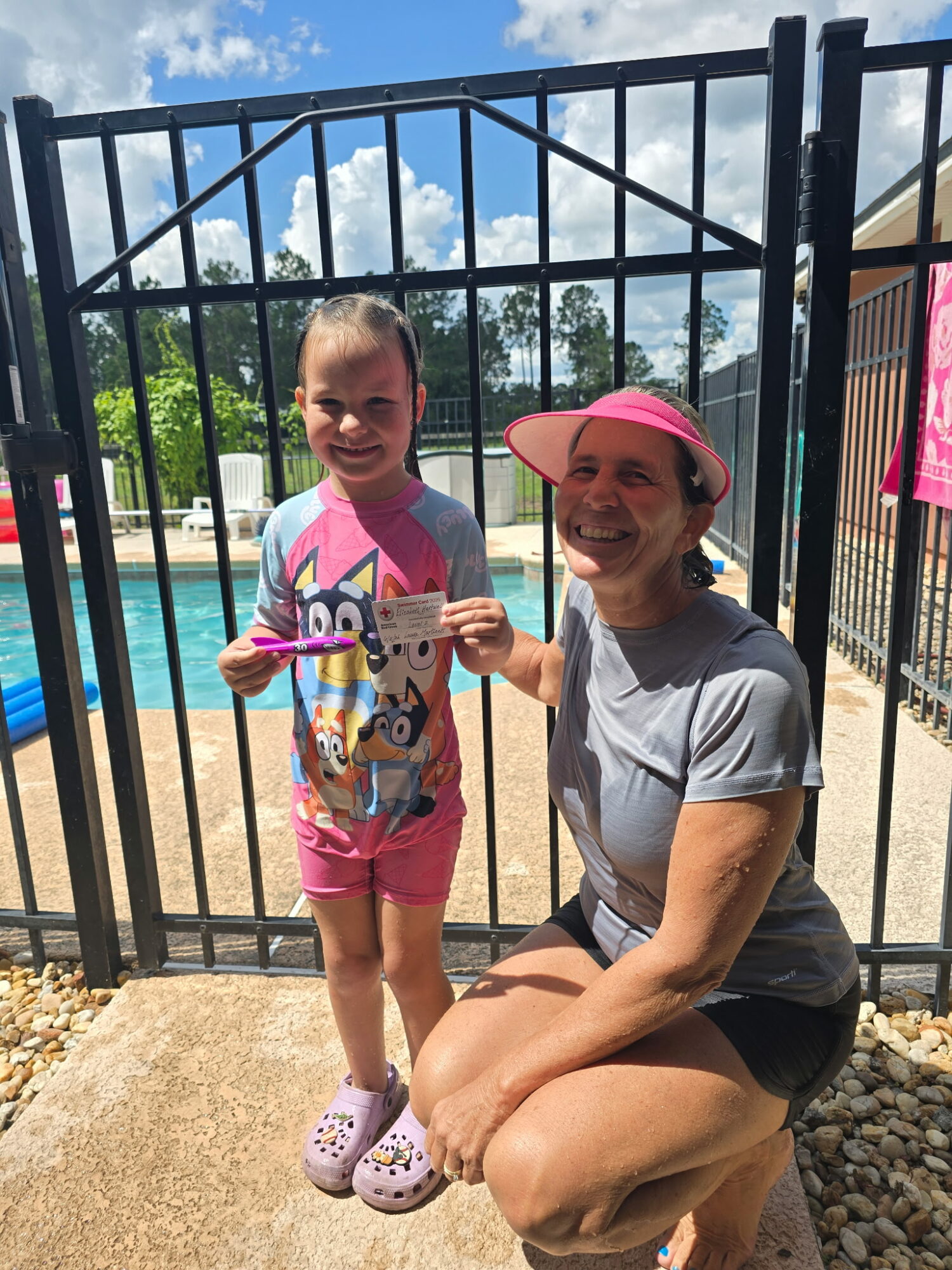 Child and woman holding a coin, standing outside near a pool with a black fence and blue sky.