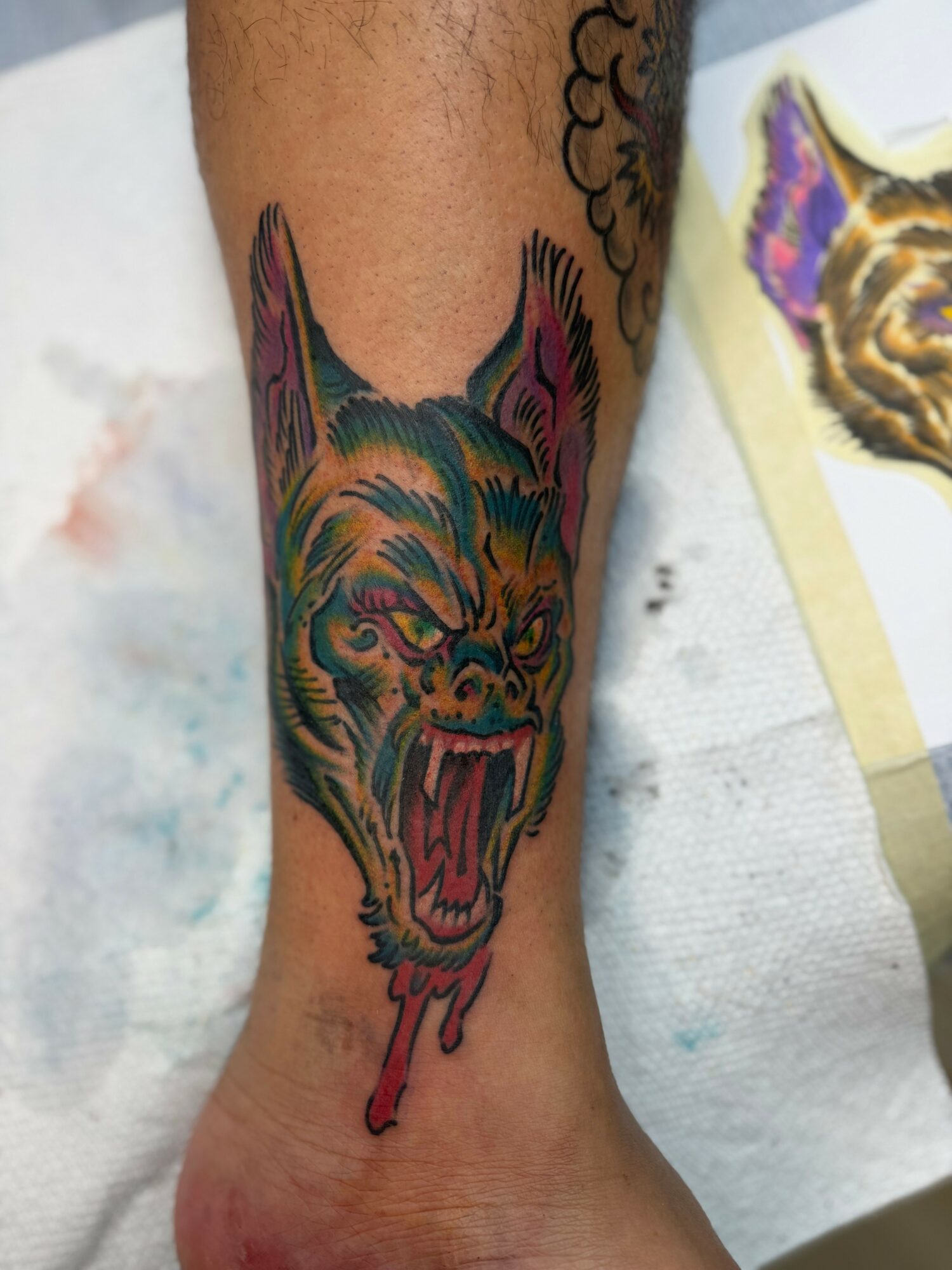 Colorful tattoo of a snarling wolf's head with dripping paint, on a person's ankle, with a cloud design nearby.