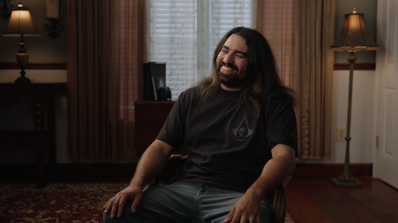 Person with long hair and beard smiling, sitting in a room with curtains and lamps, wearing a black shirt.