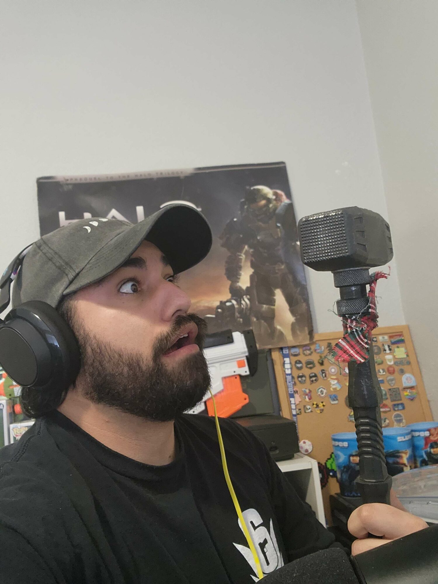 Man with headphones and cap holding a camera, with a gaming poster and shelves in background.