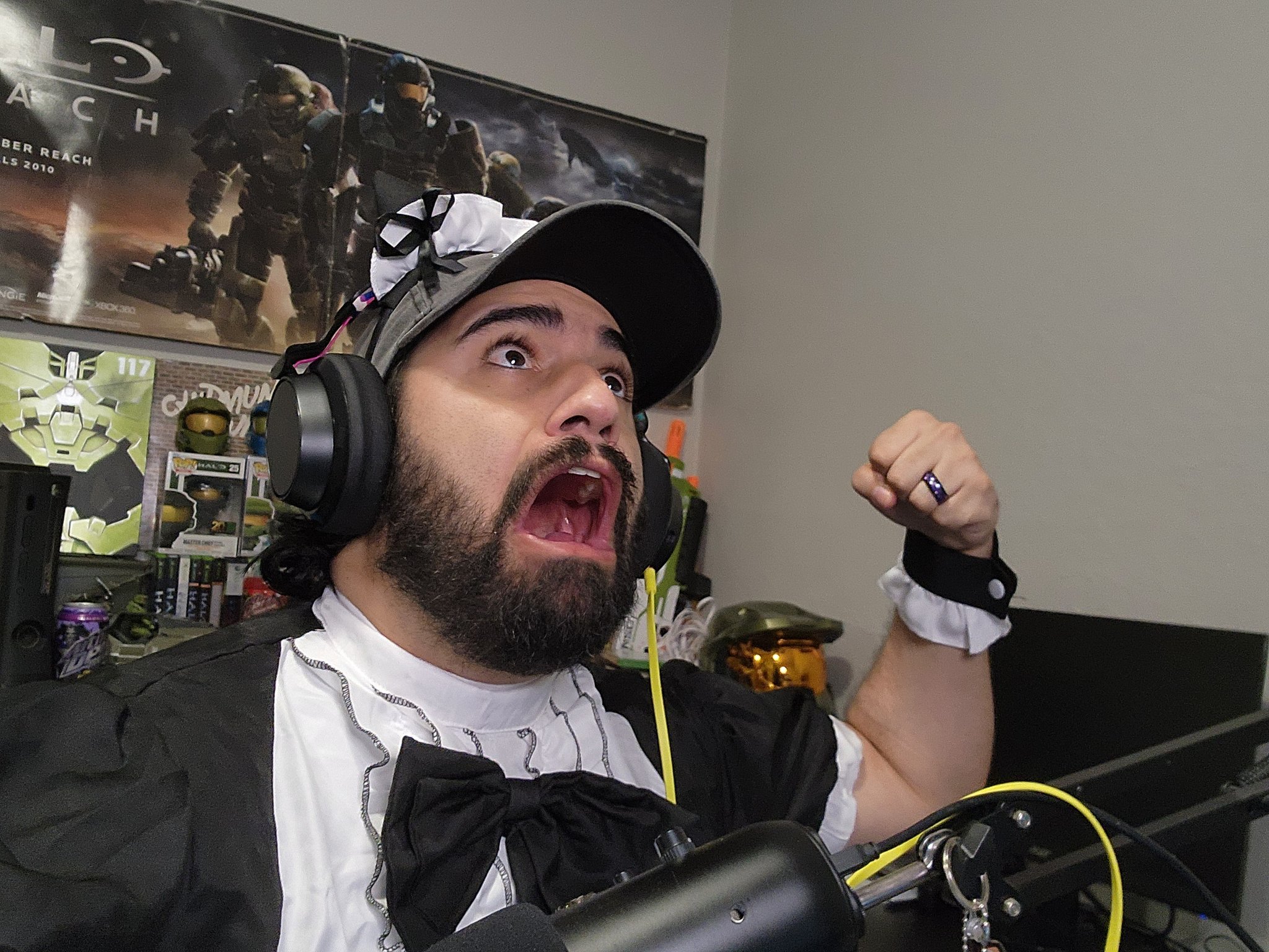 Man wearing headphones and a cap, excited and flexing his arm, in a room with posters and collectibles.