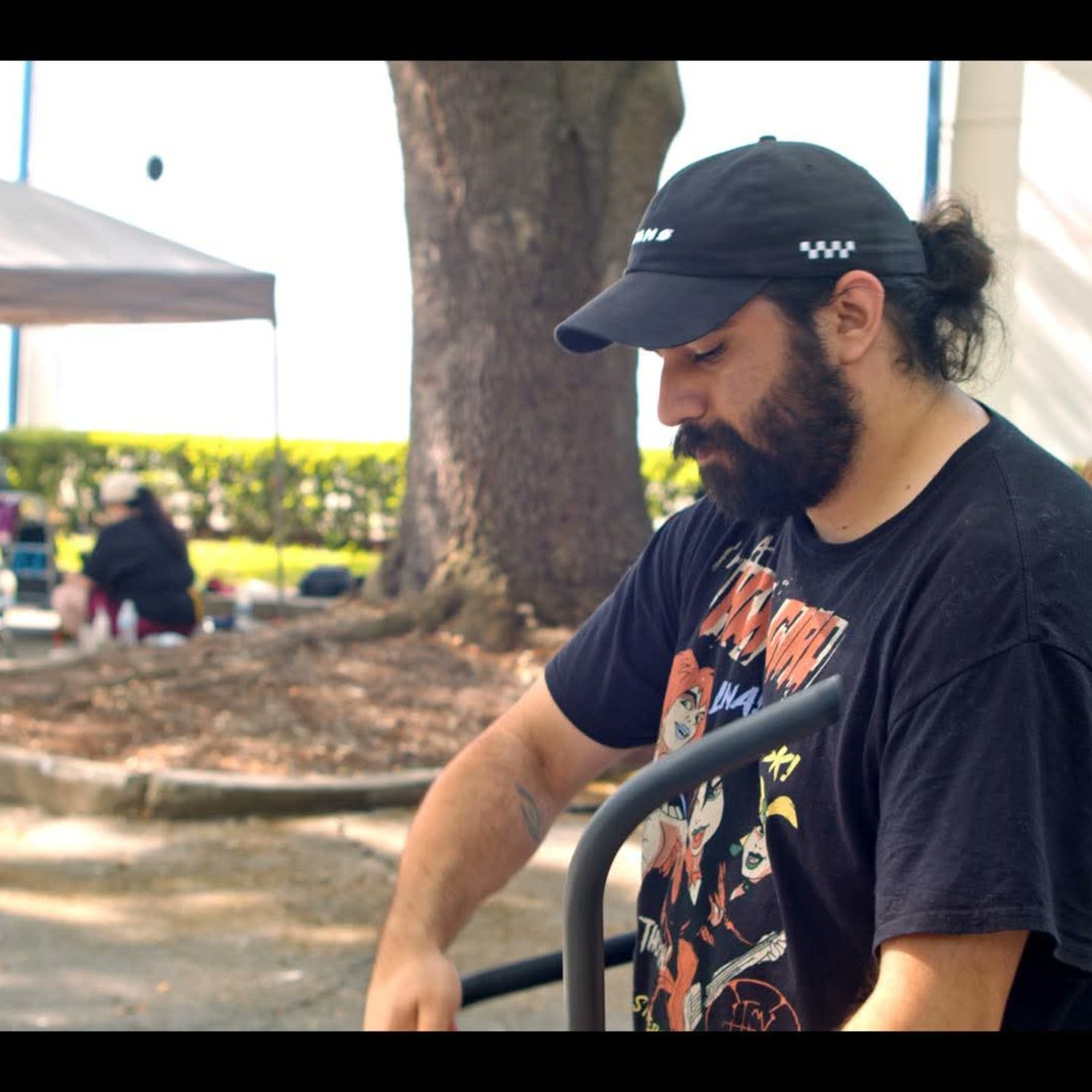 Man with dark hair and beard wearing a black cap and T-shirt, sitting outdoors near a tree.