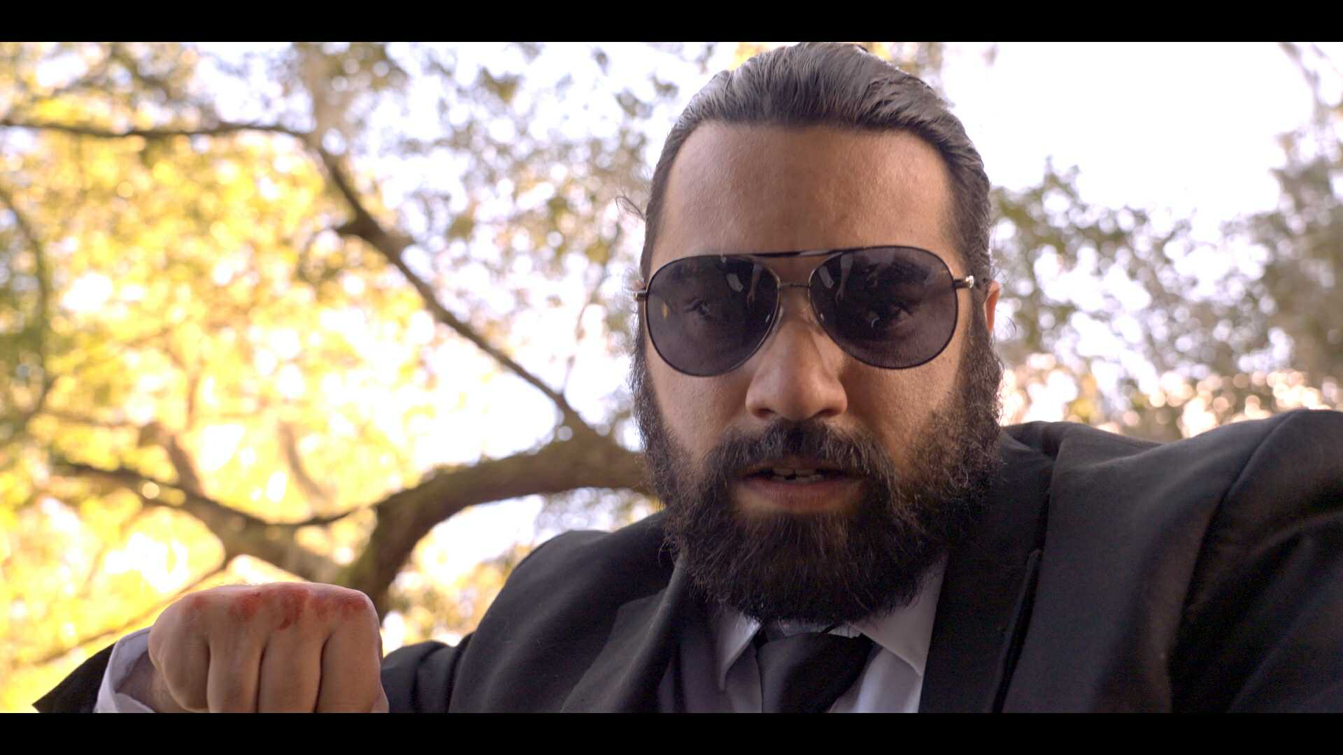 Man with dark hair, beard, sunglasses, wearing a suit outdoors with trees and sunlight in background.