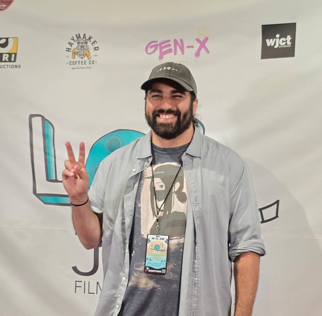 Man with beard and cap smiling and making a peace sign in front of a backdrop with logos.