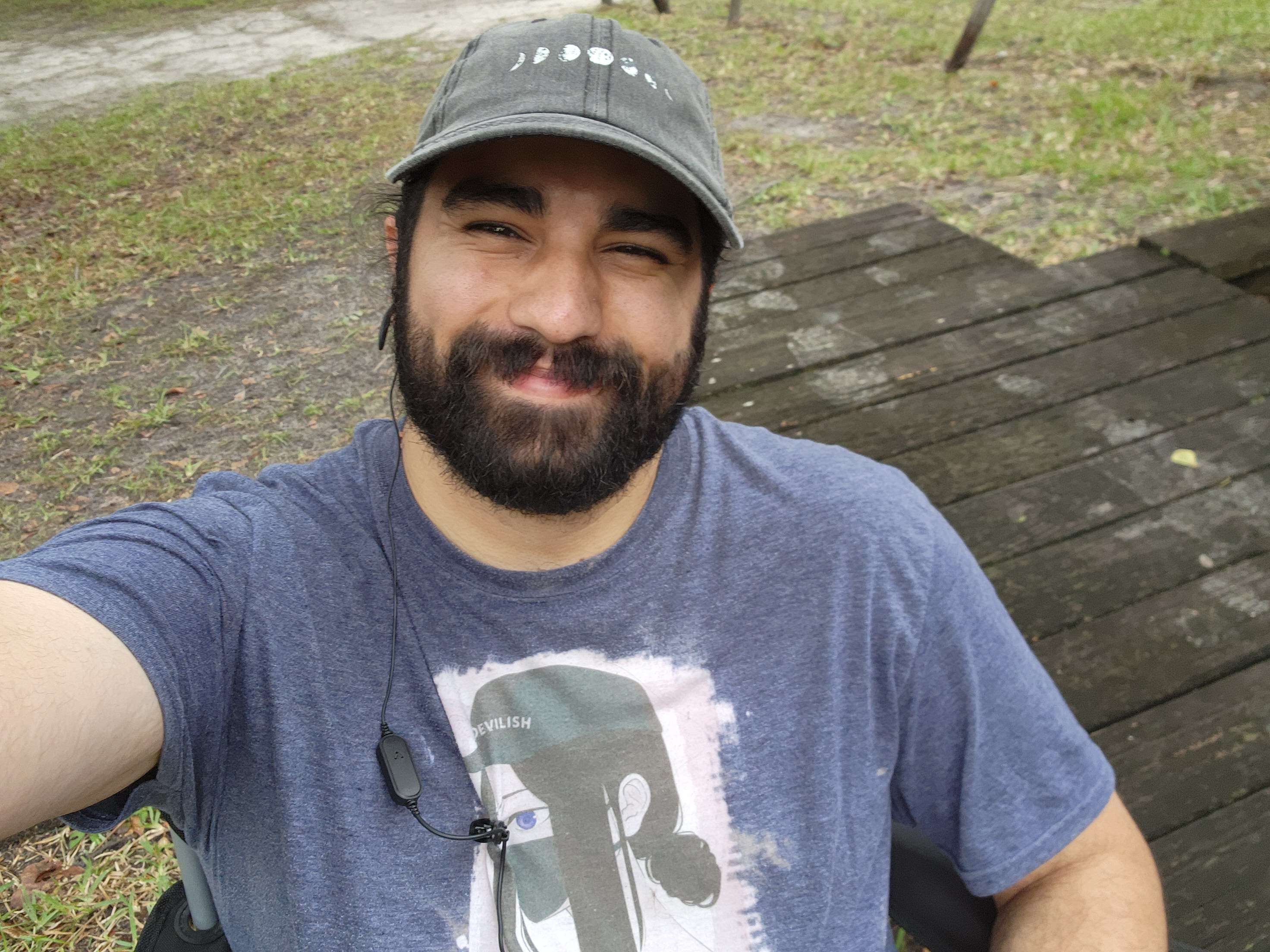 Man with a beard wearing a gray cap and blue T-shirt, smiling outdoors near wooden planks.
