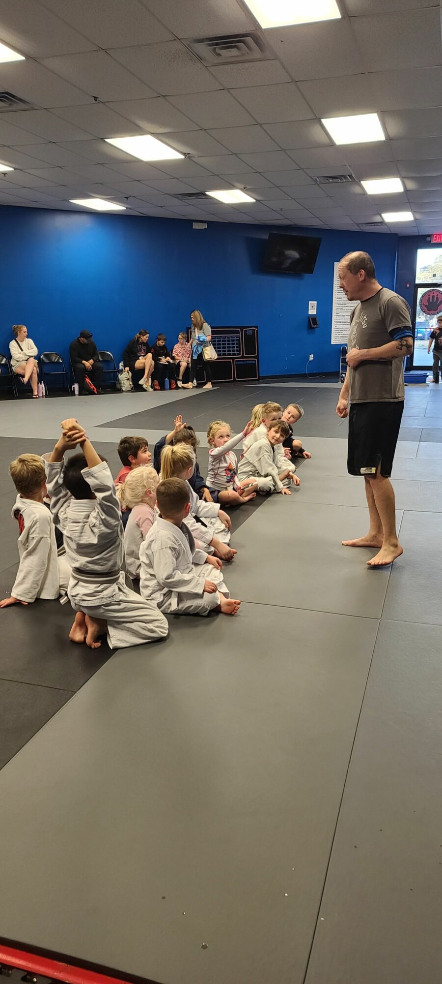 People practicing martial arts on mats in a training room with a blue wall and spectators watching.