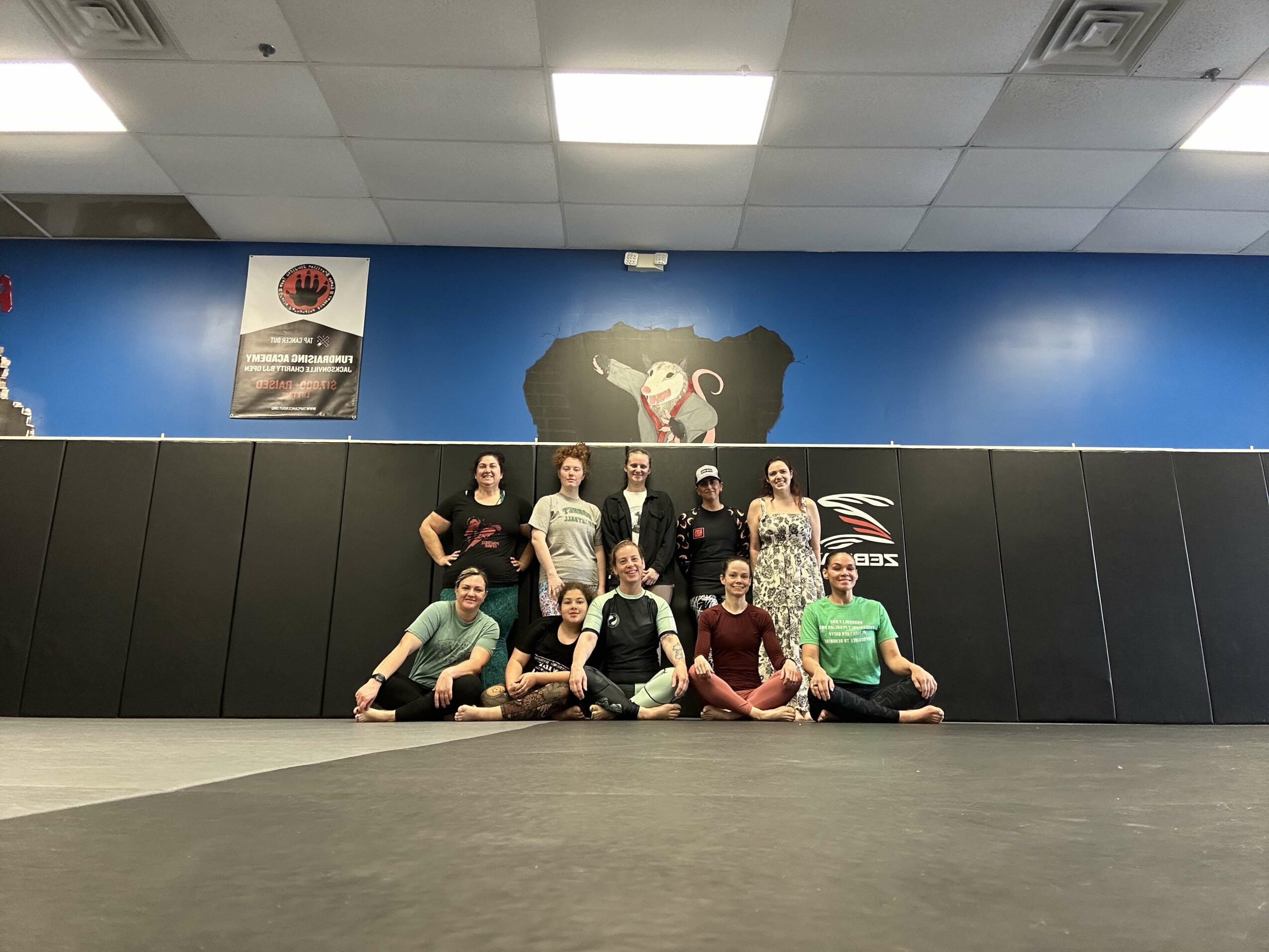 Group of nine people sitting and standing in a martial arts dojo with a blue wall and a monkey logo, ceiling lights visible.