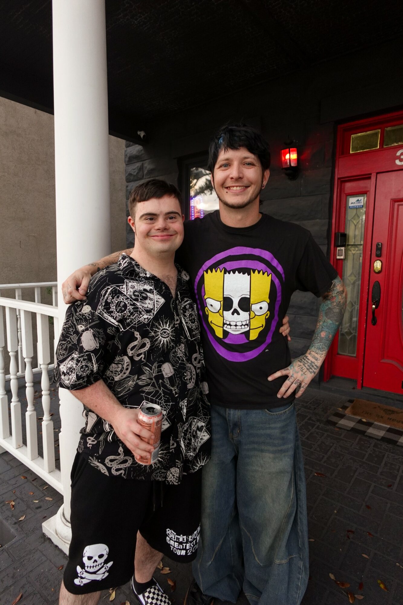 Two men standing outside near a red door, smiling, one holding a drink, one with tattoos, casual clothing.