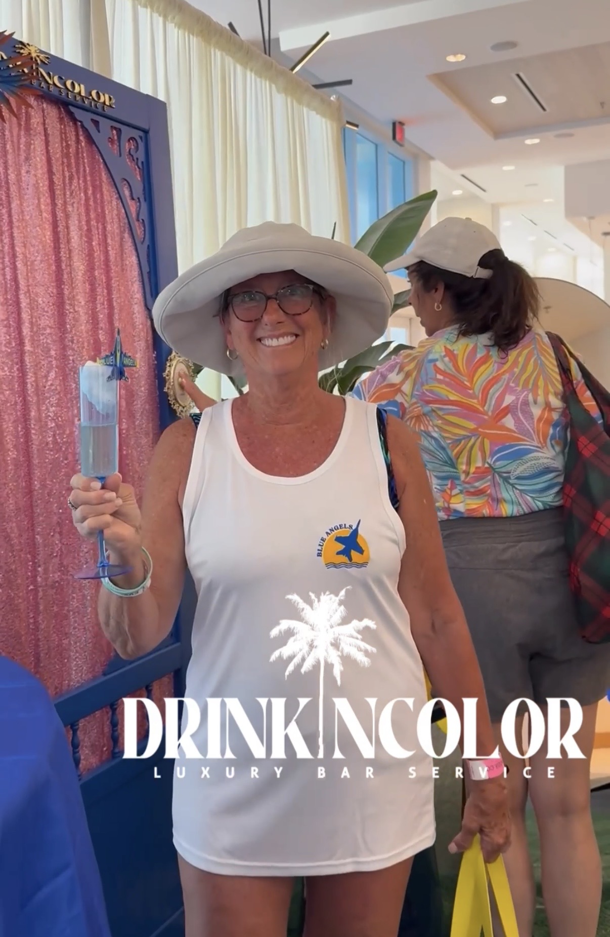 Smiling woman in white dress and large hat holding a drink at an indoor event, with other people in colorful clothing nearby.
