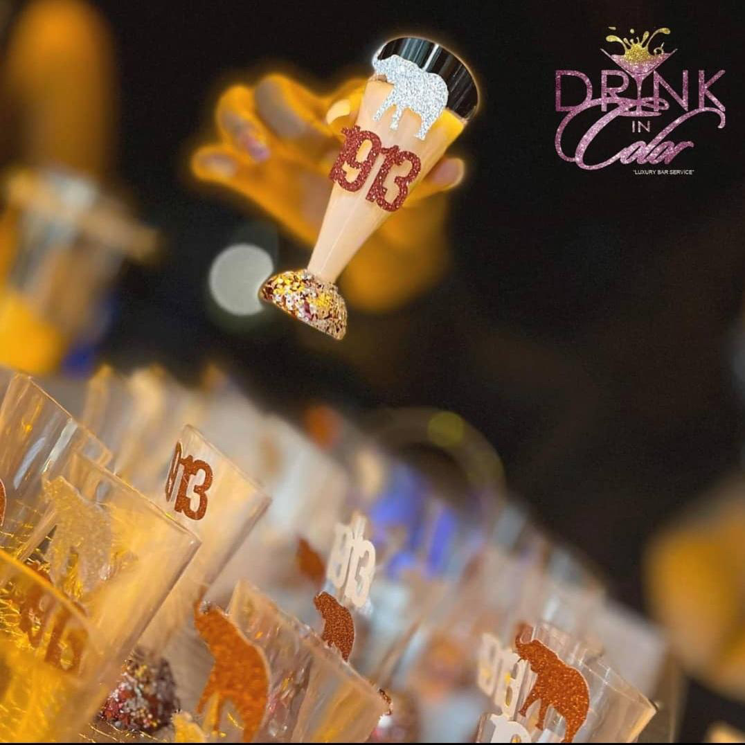 Close-up of a row of glasses with drinks, a floating beer glass decoration, and a sign reading 'Drink in' with a pink and purple design.