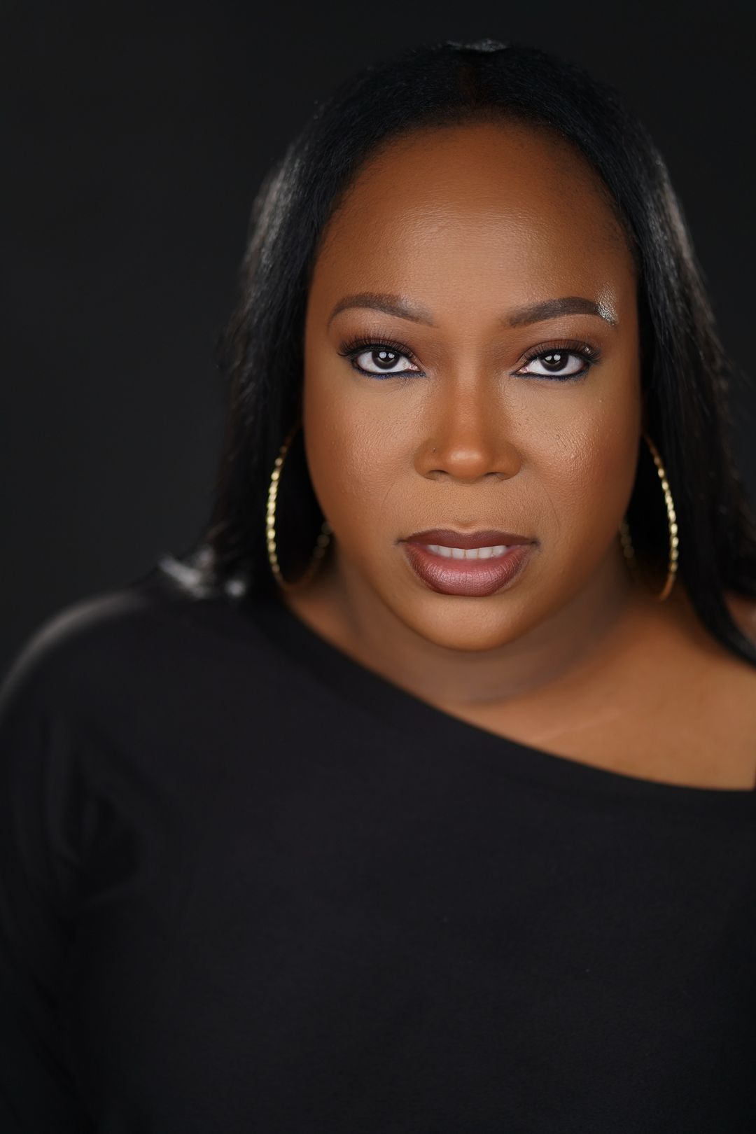 Close-up portrait of a woman with dark hair, wearing hoop earrings and a black top, looking directly at the camera.