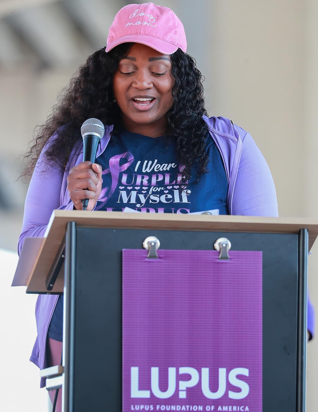 Woman speaking at a podium, wearing a pink cap and purple jacket, with a purple ribbon on her shirt, outdoors.