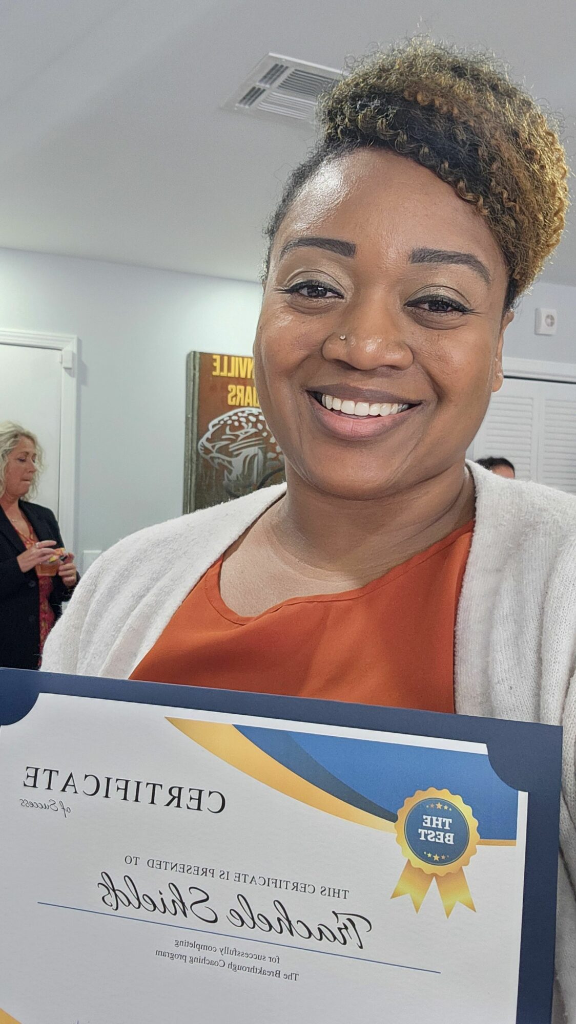 Smiling woman holding a certificate, indoors with a person in the background, white walls, and a framed picture.