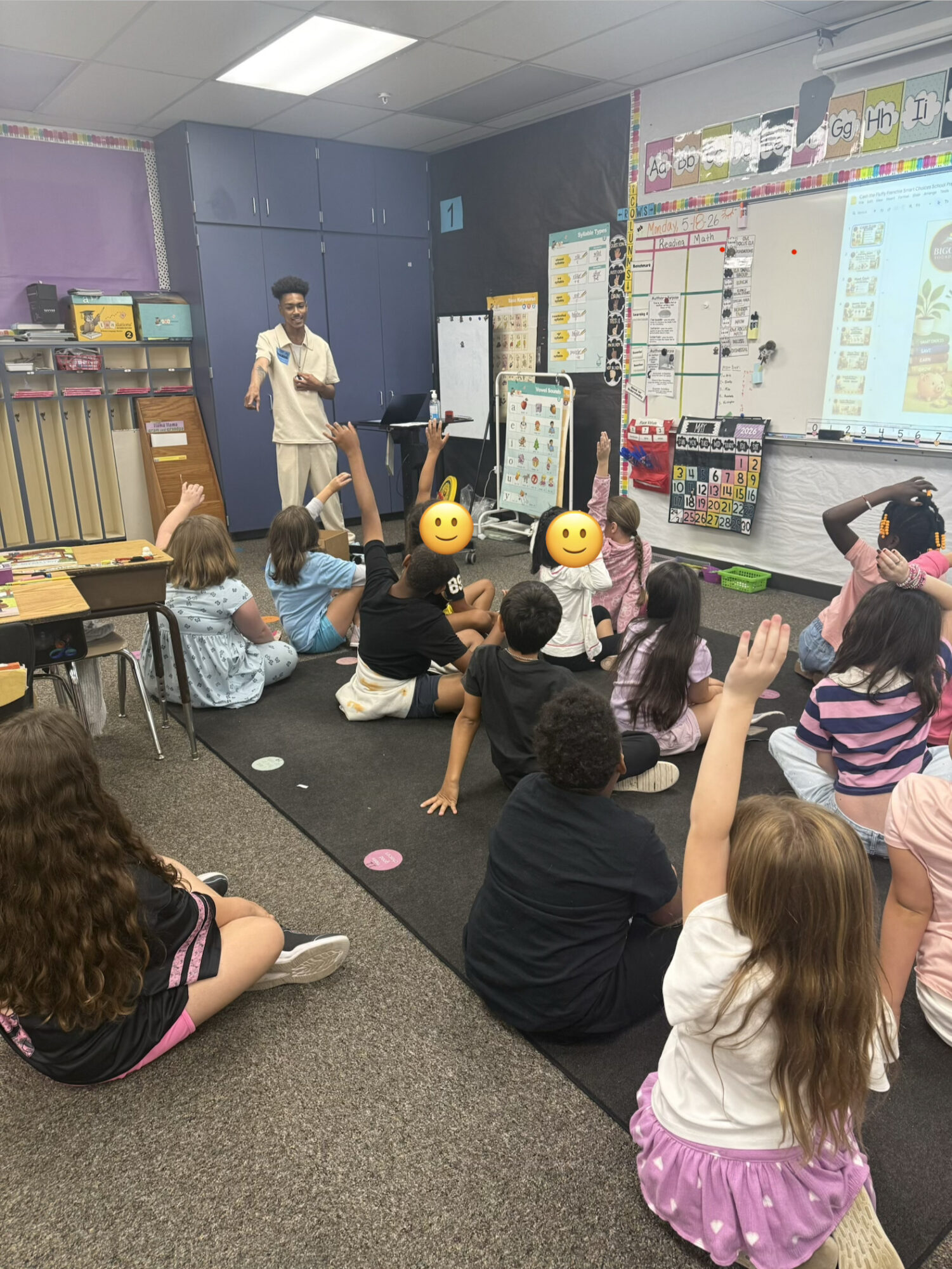 Children sitting on the floor in a classroom, raising hands, with a teacher standing near a whiteboard and a projector screen.