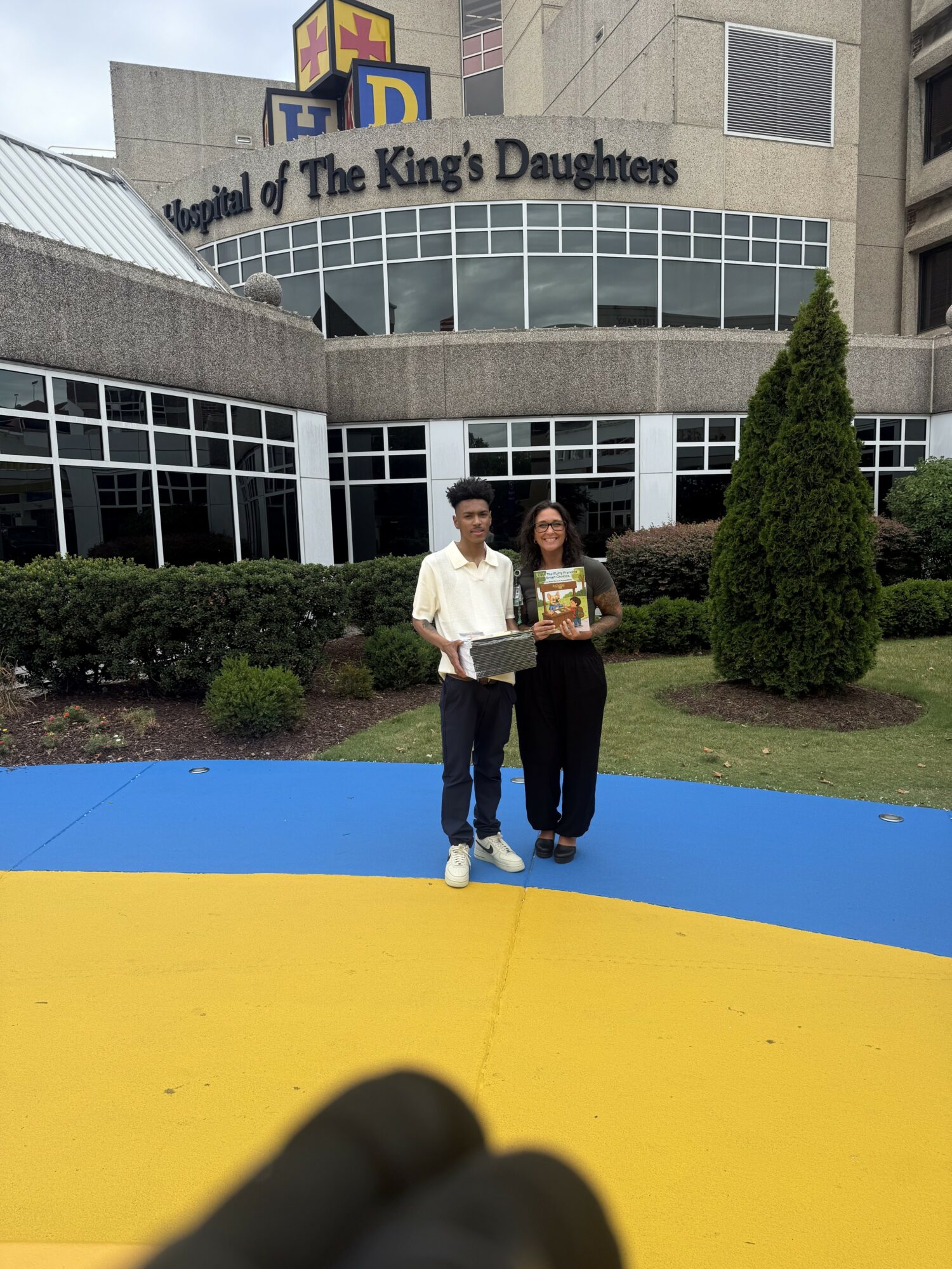 Two people stand in front of a building with a sign reading 'The King's Daughters,' holding a book or magazine, with greenery around them.