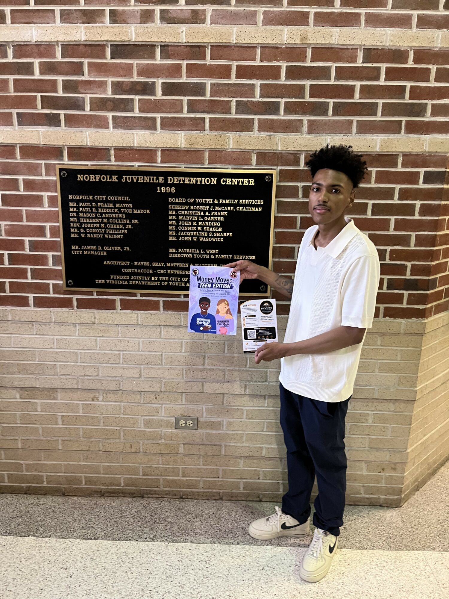 Young man with curly hair in white shirt and dark pants holding a flyer and a box, standing in front of a brick wall with a plaque.