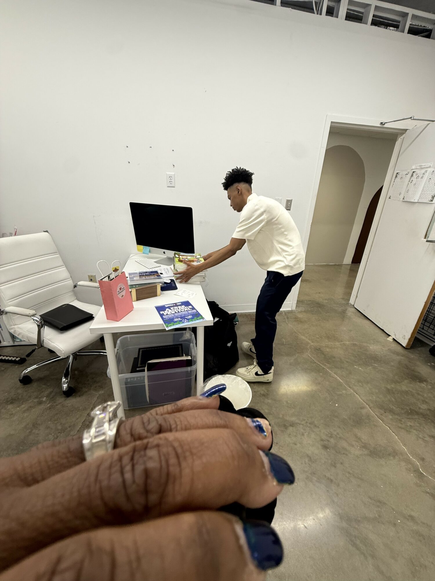 Person standing at desk with computer, office supplies, and a white chair in a modern office space.