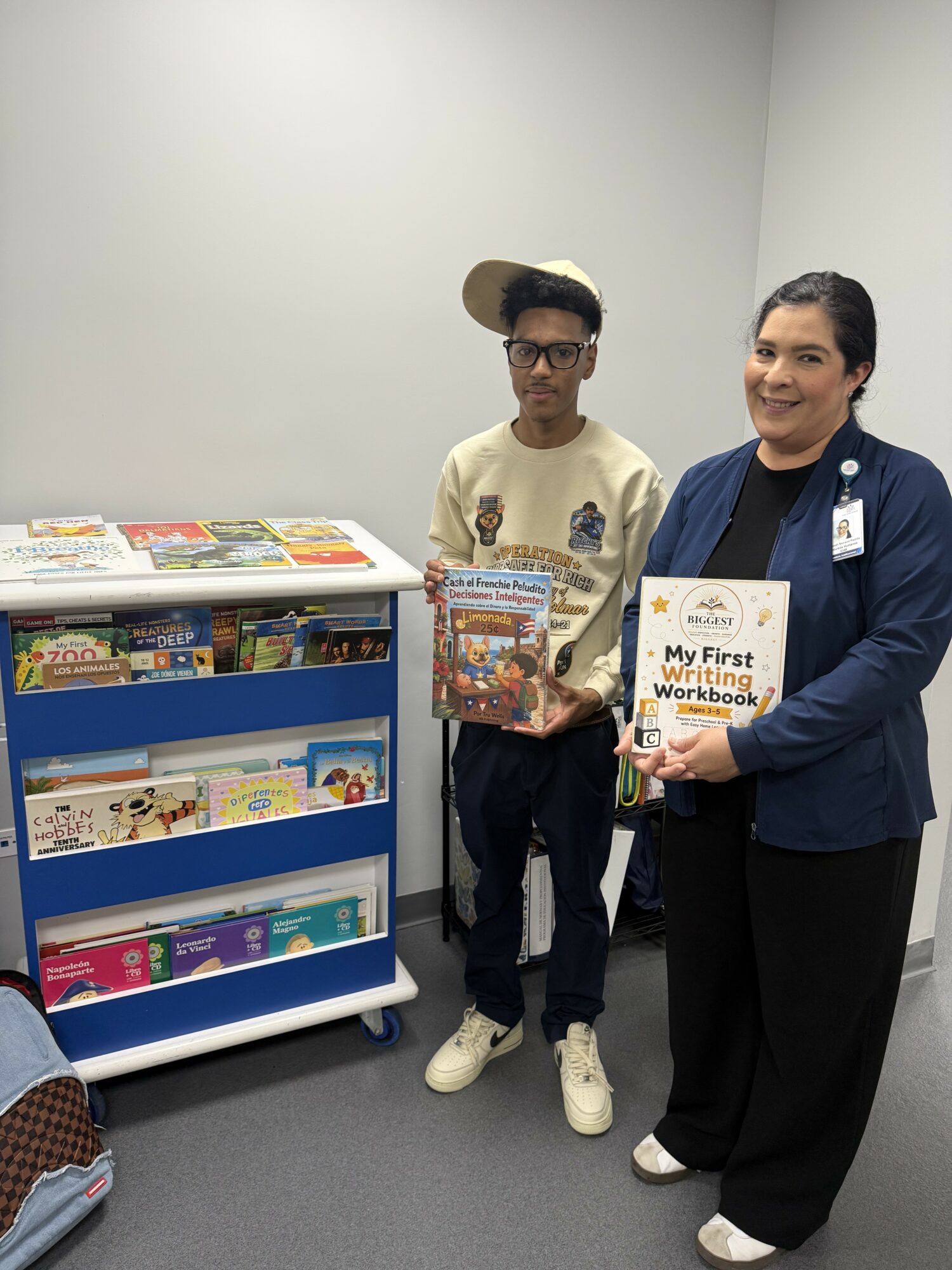 A woman and a young man stand indoors near a bookshelf with books and magazines, holding a workbook and a book, smiling.