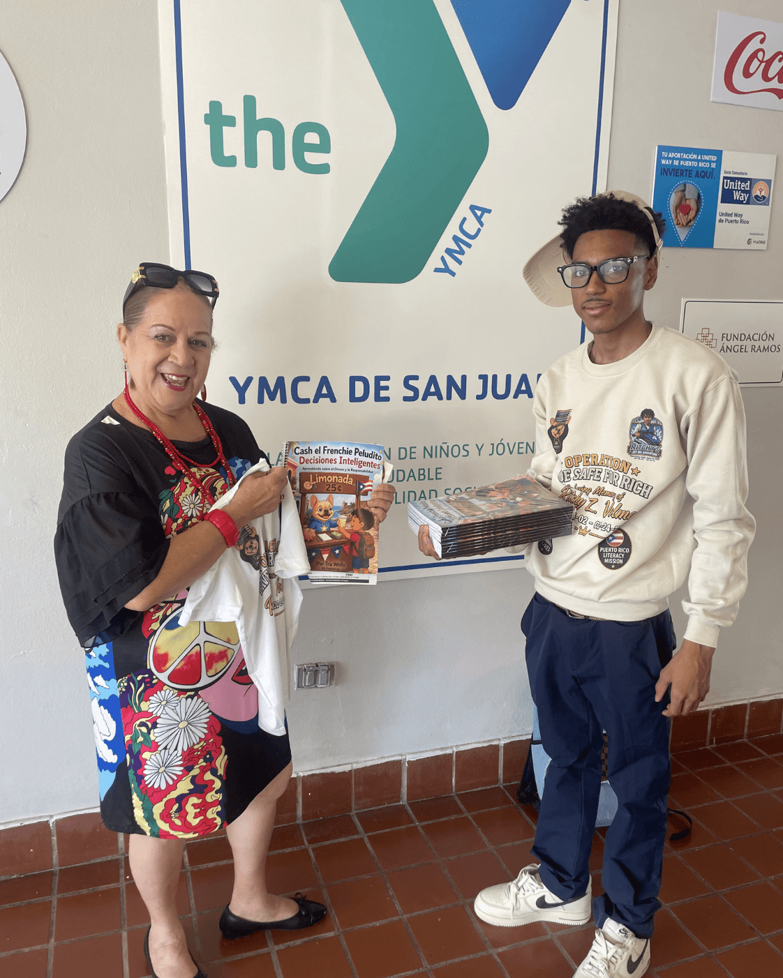 Woman and young man holding books in YMCA de San Juan, smiling, standing indoors, wall with YMCA logo behind them.