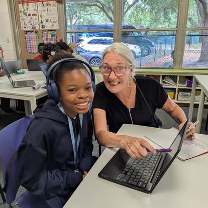 A young girl and an older woman sit at a desk with a laptop, smiling in a classroom with large windows.
