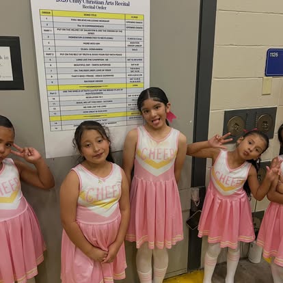 Four young girls in pink cheerleader costumes standing against a wall, smiling and posing for the camera.