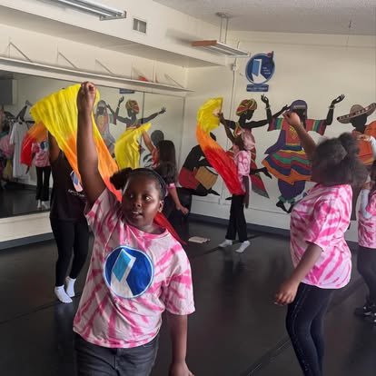 Children in pink and white shirts perform a dance with colorful scarves in a room with a mural of musicians. Group activity.