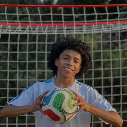 Young person with curly hair holding a colorful soccer ball in front of a goal net outdoors.