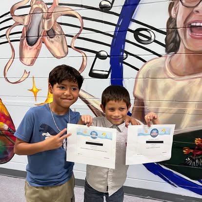 Two boys holding certificates, smiling, in front of a colorful mural with musical notes and a woman's face.
