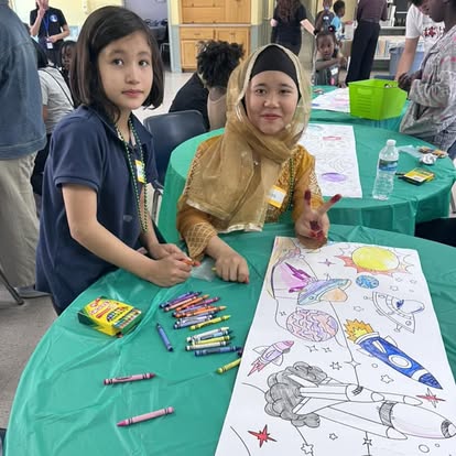 Two girls sit at a table with a large drawing of space-themed images, surrounded by markers and supplies, in a busy indoor setting.