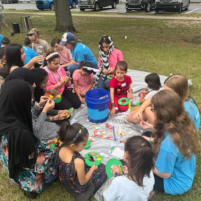 Group of children and adults gathered around a table outdoors, engaging in a craft activity with colorful materials.