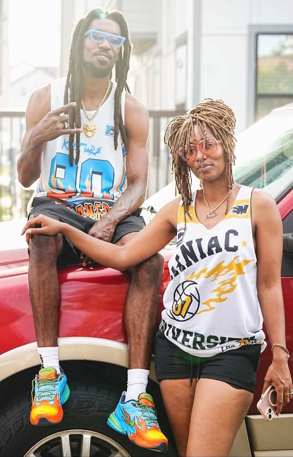 Two young people with dreadlocks and sunglasses, one sitting on a red car and the other standing, outdoors.
