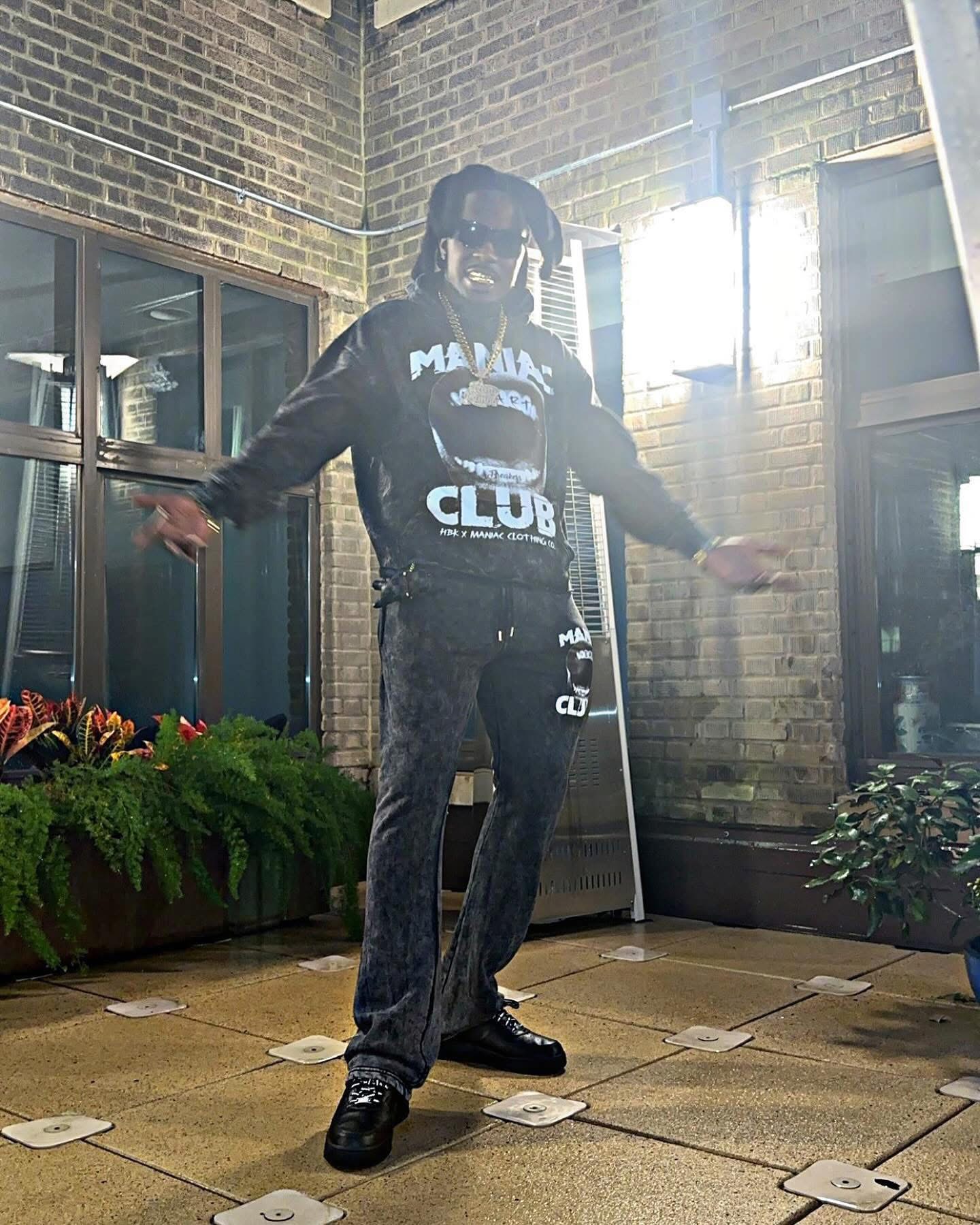 Person standing indoors with arms outstretched, wearing a black hoodie and pants, near windows and plants.