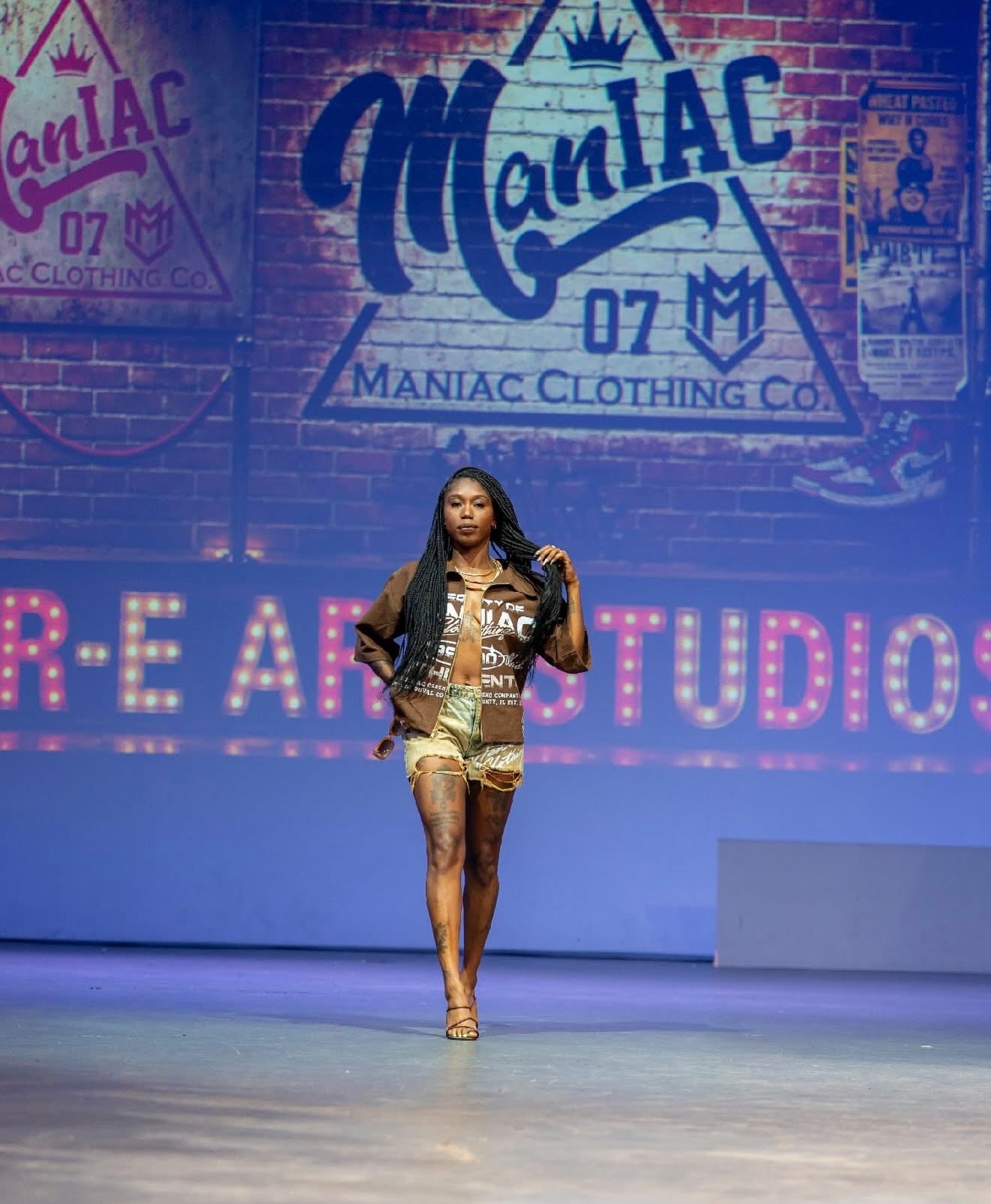 Female model walking on runway with a large sign reading 'ManiAC' and 'MANIAC CLOTHING CO.' in the background, stage lit with pink and blue lights.