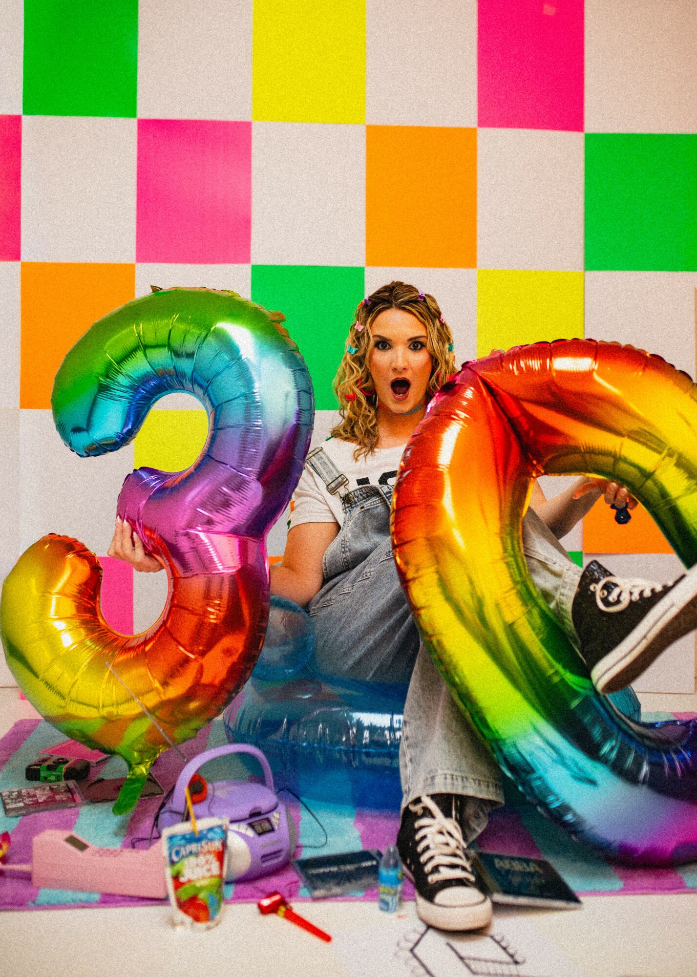 Woman sitting with colorful balloons in front of a multicolored checkered background, holding a rainbow balloon, with bags and a drink nearby.