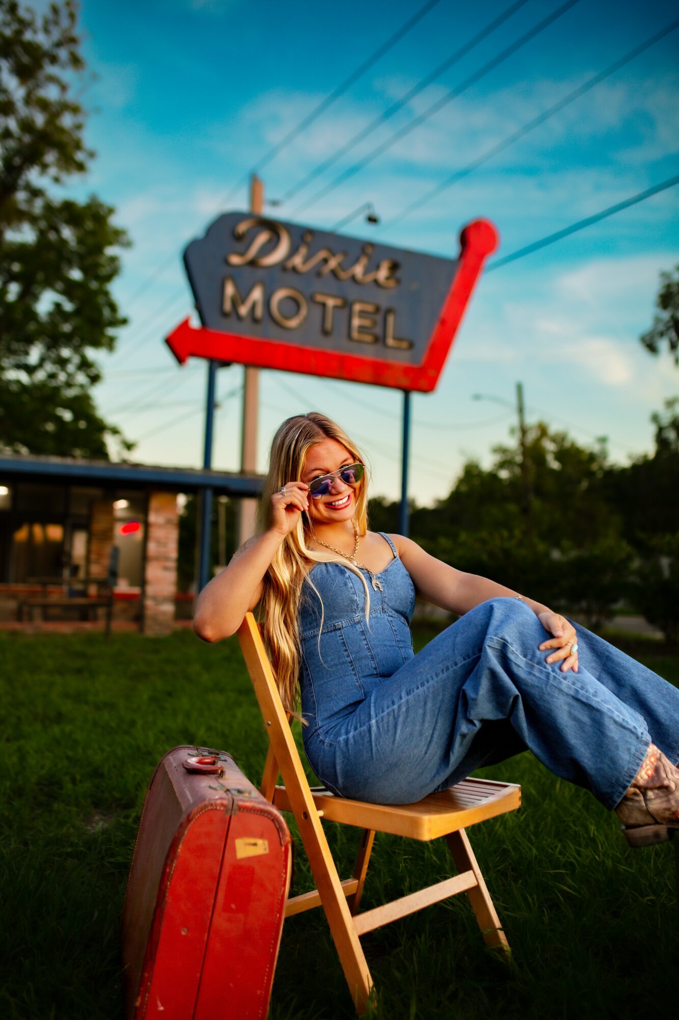 Woman in sunglasses sitting on a chair outdoors near a motel sign, holding sunglasses, with a blue sky background.