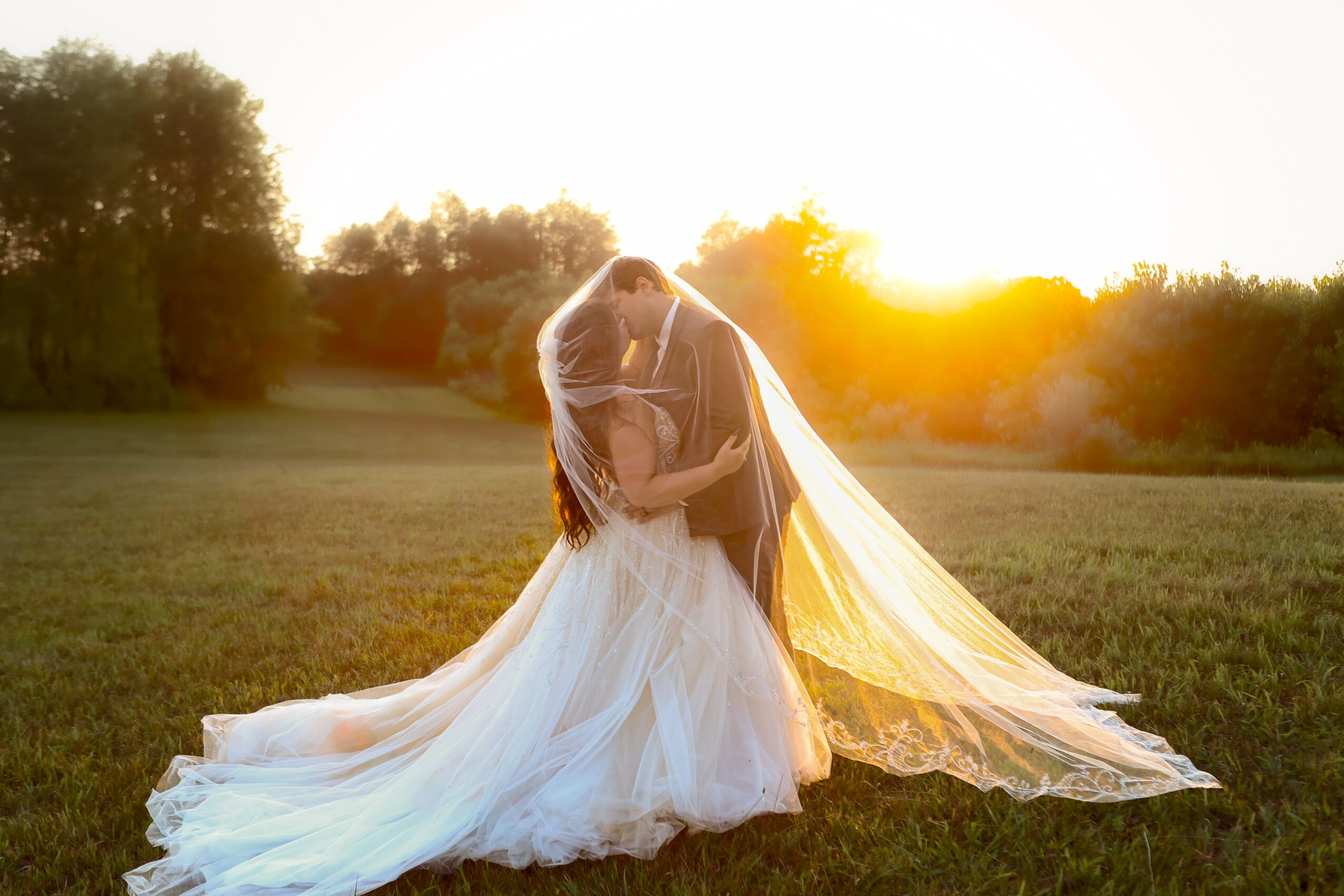Couple in wedding attire embracing outdoors during sunset, with a flowing dress and veil, in a grassy field.
