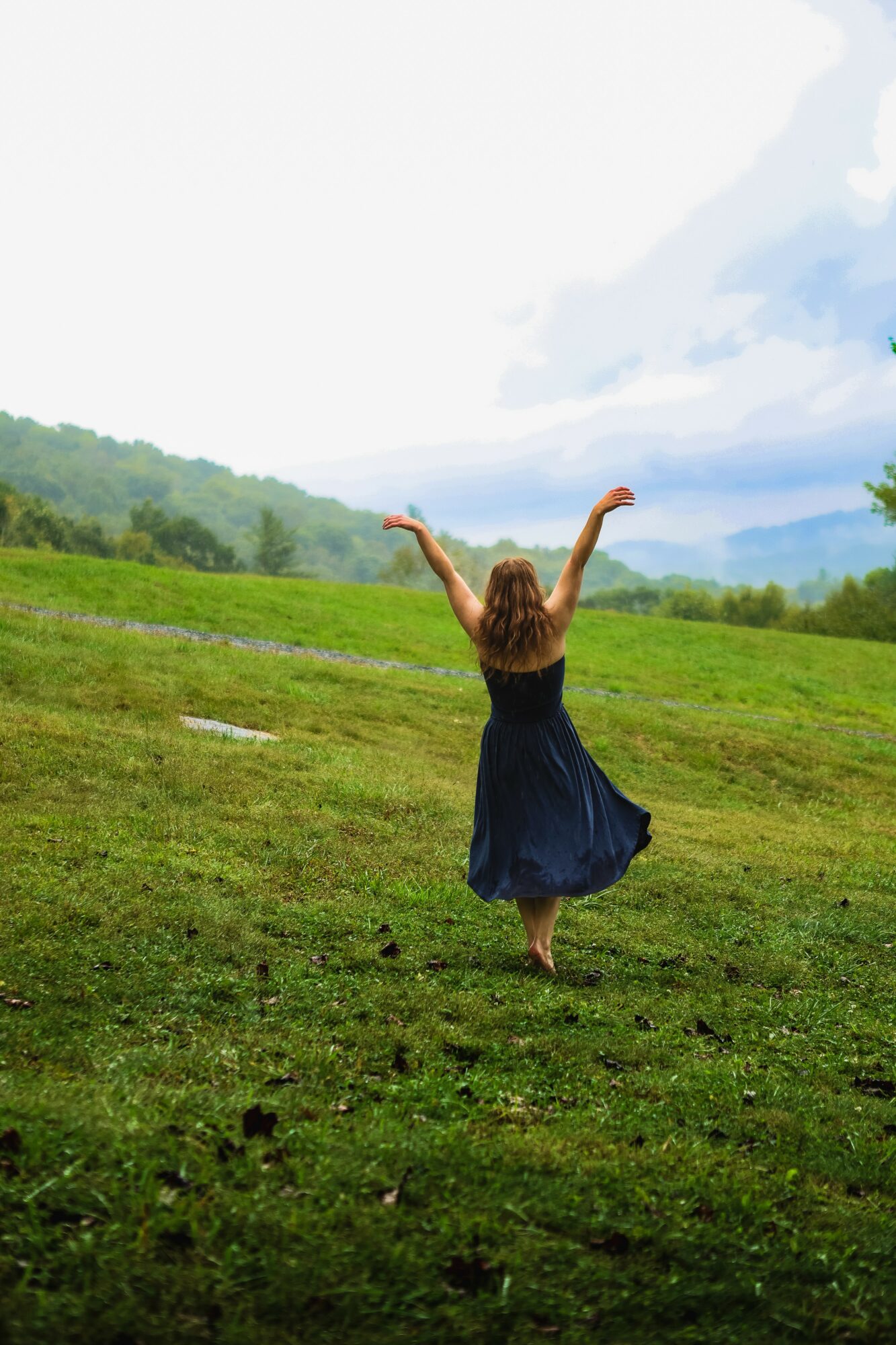 Person with long hair in a dark dress walking on grass with arms raised, landscape with trees and sky in background.