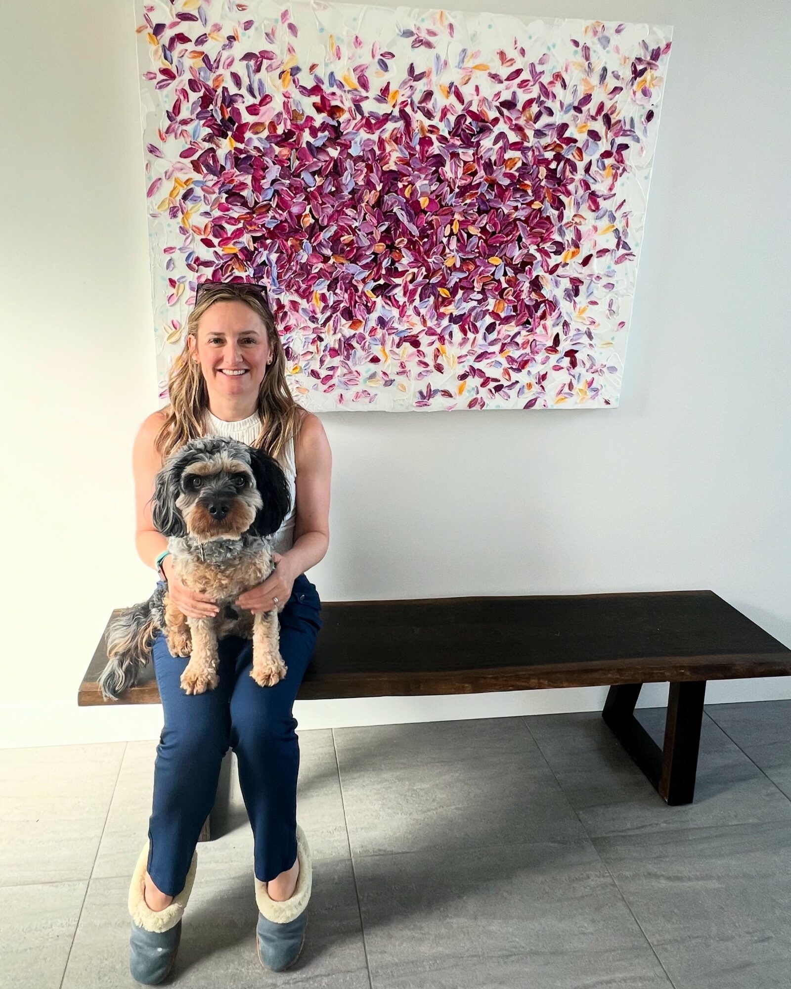 Woman holding a dog in front of a colorful abstract painting on a white wall, with a dark wooden bench nearby.
