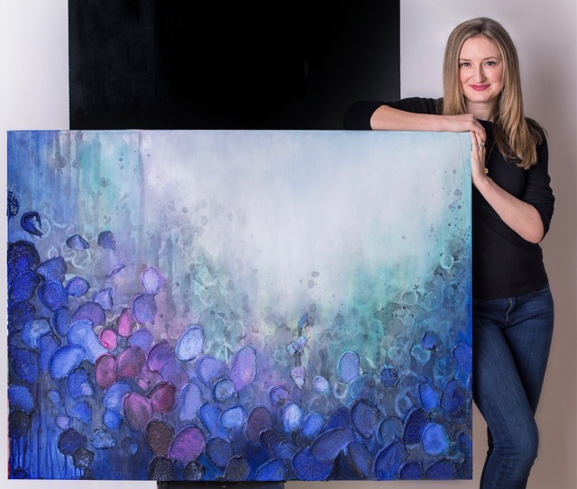 Woman with long blonde hair holding a large abstract painting with blue, purple, and green colors, smiling, standing indoors.